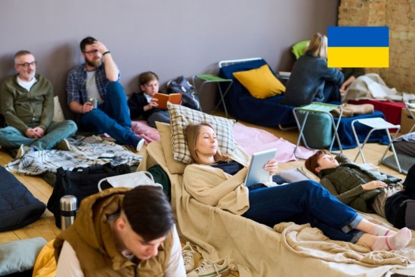 Кілька людей відпочивають на матрацах і ковдрах у приміщенні тимчасового притулку, дехто читає або користується планшетами.