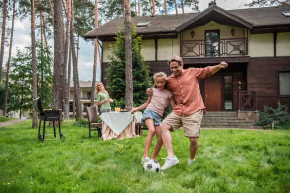 Familie im Garten vor einem Haus: Vater spielt mit Tochter Fußball, Mutter steht am gedeckten Tisch neben einem Grill.