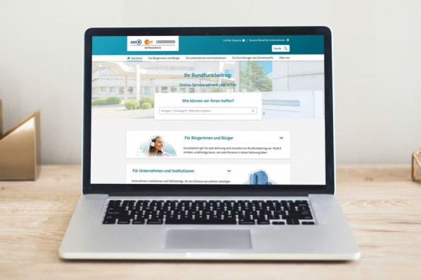 Ein Laptop auf einem Schreibtisch zeigt die Startseite der Beitragsservice-Webseite mit Suchfeld und Servicebereichen.