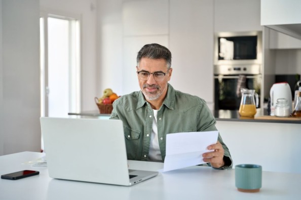 Mann mittleren Alters mit Brille und graumeliertem Haar sitzt in moderner Küche am Laptop und hält ein Dokument in der Hand.