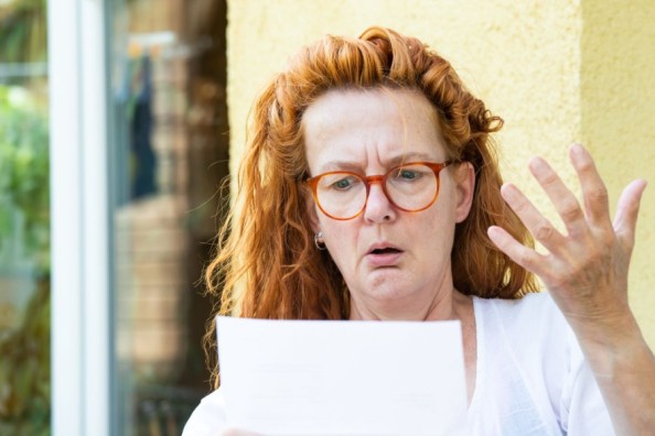 Frau mit roten Haaren und orangefarbener Brille schaut draußen überrascht auf ein Dokument und hebt fragend die Hand.