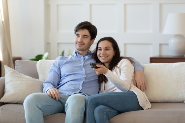 Junges Paar sitzt entspannt auf dem Sofa, schaut fern und lächelt, während die Frau die Fernbedienung hält.