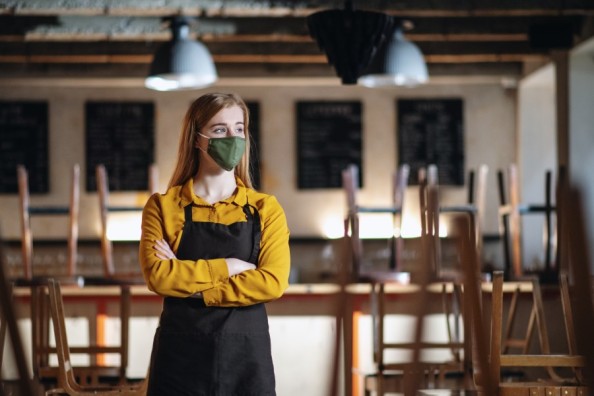 Frau mit Maske und Schürze steht mit verschränkten Armen in einem geschlossenen Café zwischen hochgestellten Stühlen.