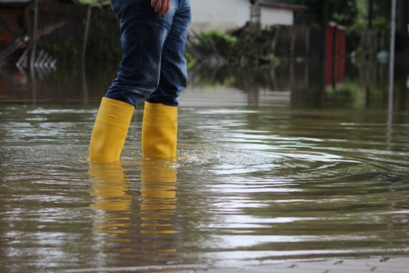 Person steht mit gelben Gummistiefeln in kniehohem Hochwasser auf einer überfluteten Straße.