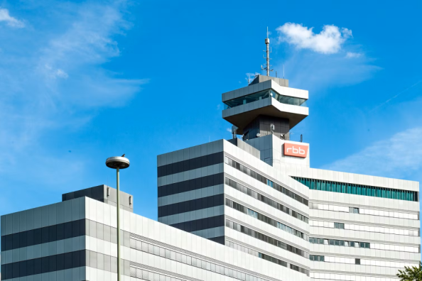 Modernes Bürogebäude mit rbb-Logo und markantem Sendeturm vor blauem Himmel.