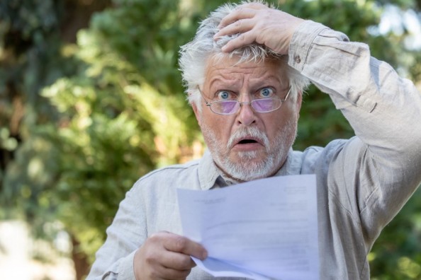 Älterer Mann mit Brille blickt erschrocken auf ein Schreiben und fasst sich mit der Hand an den Kopf.