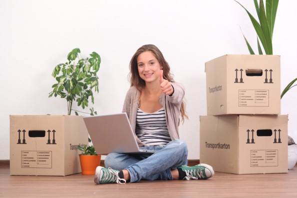 Junge Frau sitzt zwischen Umzugskartons mit Laptop auf dem Schoß und zeigt lächelnd den Daumen nach oben.