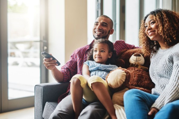 Lächelnde Familie sitzt gemeinsam mit Kind und Teddybär auf dem Sofa und schaut fern.